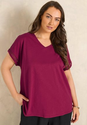 Kvinde med langt brunt hår iført en løstsiddende magentarød V-halsbluse med blonder på ærmerne og sorte bukser, der står mod en ensfarvet baggrund.