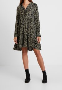 Šaty s dlouhým rukávem se zvířecím potiskem v olivově zelené barvě s černými skvrnami, s knoflíkovou légou vpředu a vícestupňovou sukní. Nosit je k nim černé kotníkové boty.