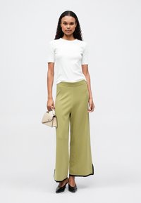 Biała prążkowana bluzka z krótkim rękawem w połączeniu z oliwkowo-zielonymi spodniami o szerokich nogawkach z wysokim stanem, z czarnymi akcentami po bokach i czarnymi płaskimi butami; trzymając małą beżową torebkę.