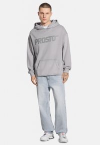Szara bluza z kapturem z napisem "PROSTO" wypisanym dużymi literami, z przednią kieszenią, noszona z jasnoniebieskimi spodniami o szerokich nogawkach.
