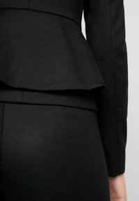 Zwart op maat gemaakt jasje met een gestructureerd oppervlak, voorzien van een peplum taille-detail en gestructureerde schouders, weergegeven van de achterkant.