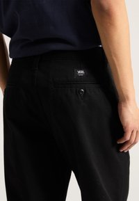 Black Vans pants feature a button and zip closure, back pockets, and a label. The fabric is smooth with a slight sheen.