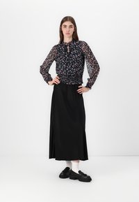 Gonna lunga nera abbinata a una blusa floreale trasparente con maniche a sbuffo e collo a fiocco. Il modello è in piedi con le mani sui fianchi, indossando scarpe nere.