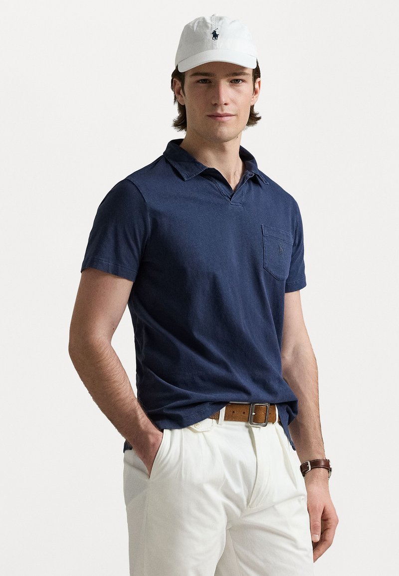 Polo-shirt bleu marine à manches courtes avec une poche, fabriqué en tissu doux. Associé à un pantalon beige clair et une ceinture brune, porté avec une casquette blanche.