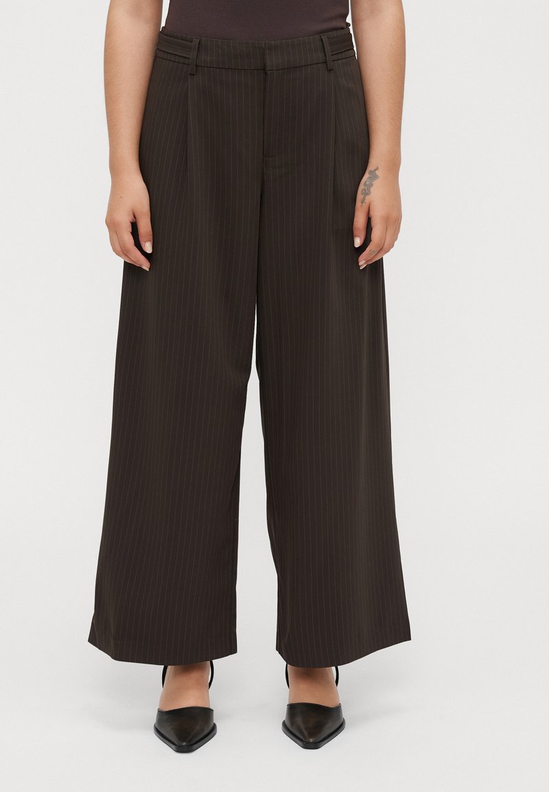 Person wearing high-waisted, wide-leg dark brown pinstripe trousers and black pointed-toe shoes, with a small tattoo on their right hand.