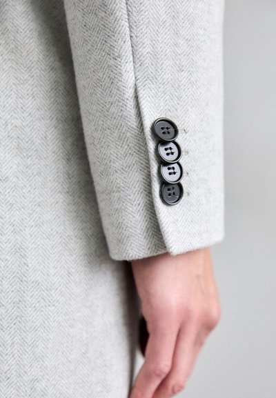 Gray herringbone jacket sleeve features four black buttons, smooth texture, and a tailored cut, highlighting the contrast between materials.