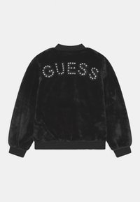 Sort faux fur bomberjakke med ribbede ærmer og kant, prydet med "GUESS" stavet med perle-lignende nitter over ryggen.