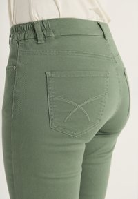 Vihreät korkeavyötäröiset housut venyvästä kankaasta, jossa on kiinnitetty vyötärö, takataskut ja kontrastiväriset saumurinteet.