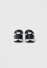 Sorte og hvide Puma-sneakers set fra bagfra, der viser logoet og strukturen på sålerne mod en ensfarvet baggrund.