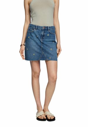 Žena oblečená v modré džínové sukni s malými žlutými květinovými vzory, béžovém topu bez rukávů, smetanových sandálech a zlatých náramcích na levém zápěstí.