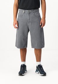 Shorts en denim gris avec une coupe décontractée, dotés de poches latérales et d'une ceinture fermée par un bouton en métal. Portés avec des baskets navy et des chaussettes blanches.