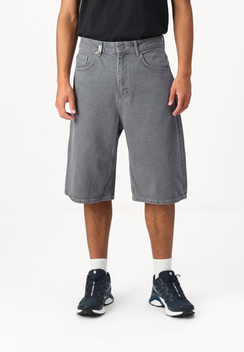 Shorts en denim gris avec une coupe décontractée, dotés de poches latérales et d'une ceinture fermée par un bouton en métal. Portés avec des baskets navy et des chaussettes blanches.