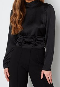 Top lungo a maniche corte in satin nero con collo alto e dettagli arricciati in vita, caratterizzato da una texture liscia e un design aderente.