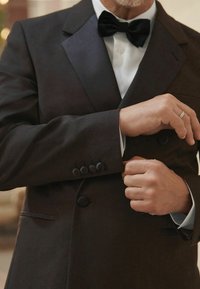 Tuxedo nero con rever in satin, maniche a quattro bottoni, e una camicia bianca con polsini blu visibili. Il papillon completa l'aspetto formale.