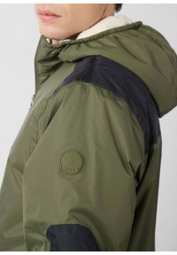 Olijfgroene jas met zwarte accenten, een capuchon en een zachte, crèmekleurige voering. Voorzien van een ronde JOTT-logo op de schouder.