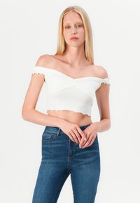 Top alb off-shoulder, scurt, cu detaliu răsucit în față, margini volan și mâneci scurte, asortat cu blugi cu talie înaltă, albaștri.