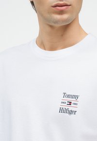 Άνδρας που φοράει απλό λευκό t-shirt με στρογγυλή λαιμόκοψη και μικρό λογότυπο "Tommy Hilfiger Since 1985" καθώς και σημαία στο αριστερό στήθος.