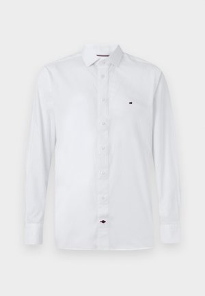 Balts garais piedurknēm pogājams krekls no gludas auduma, ar klasisku apkakli un mazu daudzkrāsainu logo izšuvumu uz krūtīm.