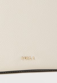 Svetlo bež teksturirana usnjena površina z majhnimi zlatimi črkami, ki na spodnjem robu tvorijo napis "FURLA".