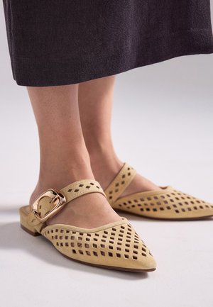 Zapatos planos beige con punta puntiaguda, con patrones cuadrados recortados y una correa ancha que presenta una gran hebilla dorada, usados con una falda oscura.