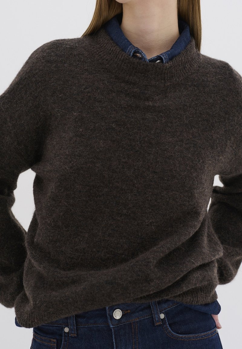 Femme portant un pull marron foncé en tissu pelucheux par-dessus une chemise bleue à col et un jean bleu foncé, les mains posées sur les hanches.