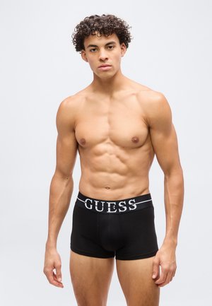 Μυώδης νεαρός με σγουρά μαλλιά που φοράει μαύρα εσώρουχα boxer Guess, στέκεται μπροστά σε ένα απλό ανοιχτό γκρίζο φόντο.