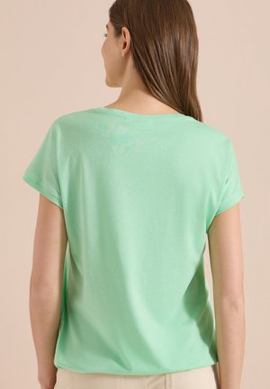 Femme aux longs cheveux bruns portant une chemise vert clair à manches courtes, vue de dos devant un fond beige uni.