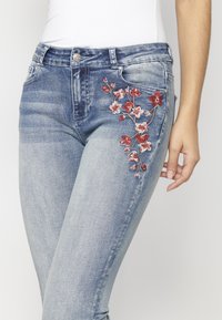 Blaue Denim-Jeans mit einem ausgewaschenen Design, versehen mit roter Blumenstickerei auf der Vordertasche. Schmal zulaufendes Bein und normale Reißverschlussverschluss.