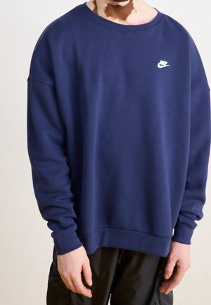 Osoba ubrana w granatową bluzę Nike z białym logo na piersi oraz czarne spodnie, stojąca na jednolitym tle.