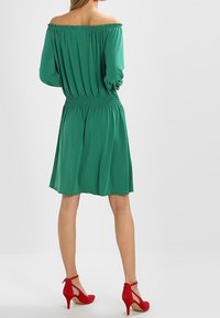 Zielona sukienka na ramiączkach z marszczonym pasem, lekko powłóczysta spódnica i długie rękawy, w połączeniu z czerwonymi szpilkami.