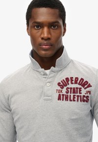 Grijs polo shirt met lange mouwen en een kraag, met op de linkerkant van de borst het bordeauxrode geborduurde logo "SUPERDRY TOK STATE JPN ATHLETICS". Zachte textuur.