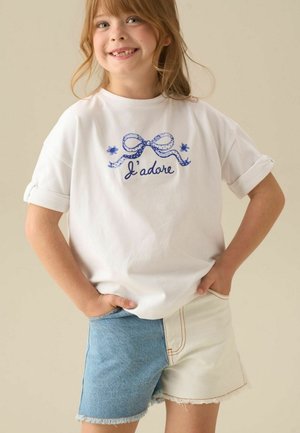 Junges Mädchen lächelt mit Händen in den Taschen, trägt ein weißes T-Shirt mit blauer Schleife und "J’adore"-Text sowie zweifarbige blau-weiße Denim-Shorts.