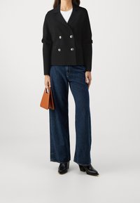 Czarna dwurzędowa marynarka z srebrnymi guzikami, zestawiona z szerokimi dżinsami w kolorze niebieskim i czarnymi botkami. W ręku brązowa torebka.