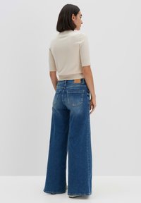 Jean en denim bleu à jambes larges avec une finition décolorée et une étiquette en cuir sur la taille. Associé à un haut ajusté de couleur crème.