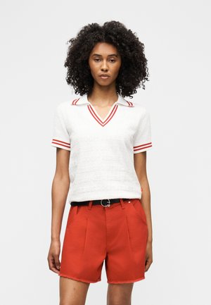 Kokkupandud juustega naine, kes kannab valget lühikest varrukatega polosärki punaste triipudega ja kõrge vöökohaga punaseid lühikesi pükse musta vöögaga.