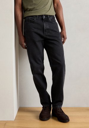 Black denim jeans with a straight-leg cut, featuring five pockets and a button closure, paired with dark brown boots on a wooden floor.