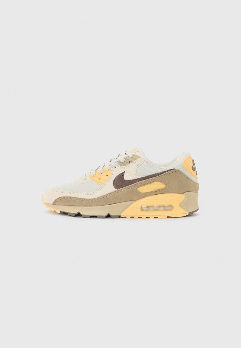 Sneaker Nike Air Max 90 cu un superior bej, accente maro, detalii galbene și o unitate de aer vizibilă în călcâi. Talpa din cauciuc completează designul.