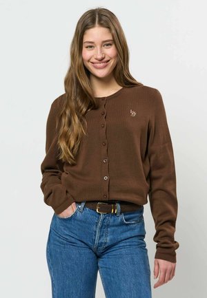 Jeune femme aux longs cheveux portant un pull marron boutonné et un jean bleu, souriant avec une main dans la poche.