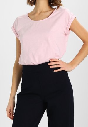 Kobieta ubrana w jasnoróżową koszulkę z krótkim rękawem, włożoną w wysokie czarne spodnie, stoi z jedną ręką na biodrze.