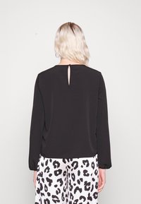 Schwarzes Langarmoberteil mit rundem Ausschnitt und einem kleinen Schlüssel-Loch-Detail hinten, kombiniert mit weißen Hosen mit einem schwarzen Leopardenmuster.
