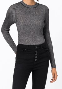 Kobieta w dopasowanym, długim rękawie, prążkowanej, metalicznej szarej bluzce wpuszczonej w czarne spodnie o wysokim stanie z pięcioma guzikami z przodu.