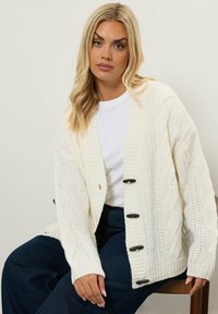 Krämfärgad kabelstickad cardigan med stora knappar, ribbade ärmslut och en lös passform, bärs över en vit T-shirt och mörka jeans.