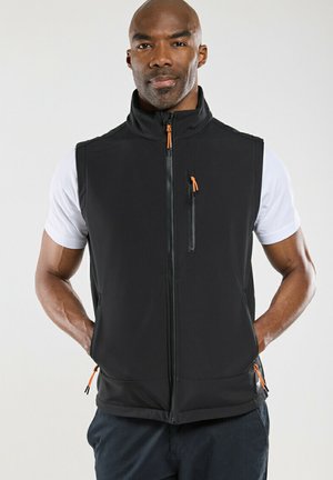 Homme portant un gilet sans manches noir à fermeture éclair avec tirettes orange sur une chemise blanche à manches courtes, debout devant un fond clair.