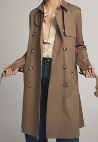 Personne portant un trench beige sur un chemisier crème et un jean bleu, tenant la ceinture du manteau avec les deux mains.