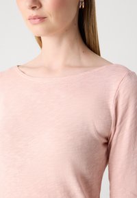 Žena u svijetlo ružičastom teksturiranom topu s brojnim okruglim izrezom i zlatnim okruglim naušnicama, prikazuje vrat i gornji dio torza naspram jednostavne pozadine.