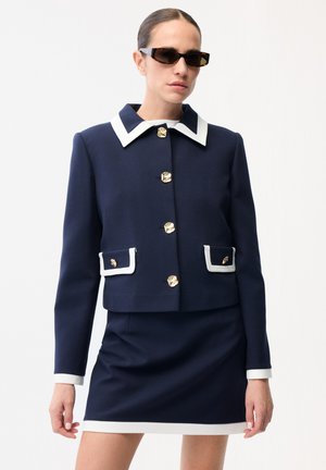 Kvinde iført marineblå skræddersyet jakke med hvide kanter og guldknapper, matchende nederdel og mørke solbriller mod en ensfarvet baggrund.