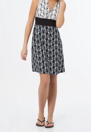 Žena v bezrukávových černobílých šatech s listovým vzorem, s černým páskem v pase a černými sandály, stojící s jednou rukou u krku.