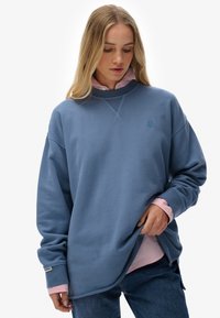 Blauwe oversized sweatshirt met een gestructureerde afwerking, voorzien van een ronde halslijn, verlaagde schouders en een geborduurd logo. Gelaagd over een roze shirt.