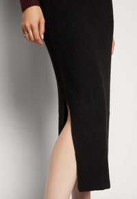 Vestido preto de malha canelada com uma fenda lateral até à coxa. O tecido tem uma textura suave, com uma silhueta justa que termina abaixo do joelho.