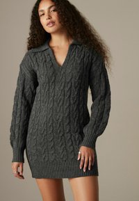 Robe pull en tricot gris avec un décolleté en V, des manches longues et un ourlet côtelé. Motif texturé sur l'ensemble, mettant en valeur des coutures épaisses.
